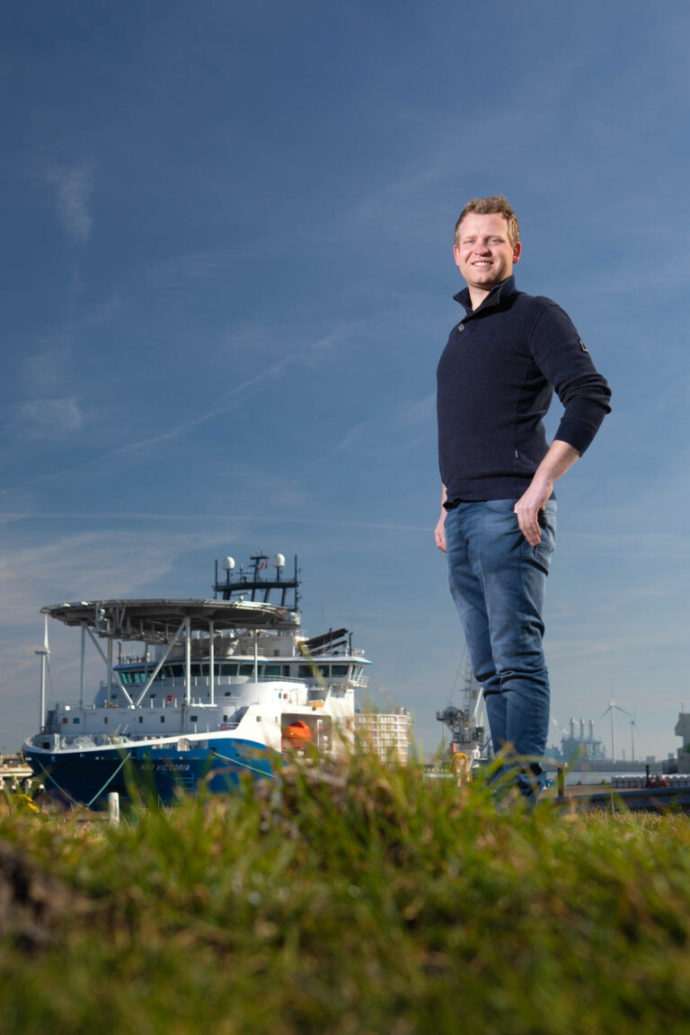 Een man staat buiten op een grasveld met een grote scheepswerf of haven op de achtergrond. Hij draagt een donkerblauwe trui en jeans, en glimlacht terwijl hij in de camera kijkt. De foto is gemaakt op een zonnige dag met een heldere blauwe lucht. Fotograaf: Jan Buwalda, Groningen.