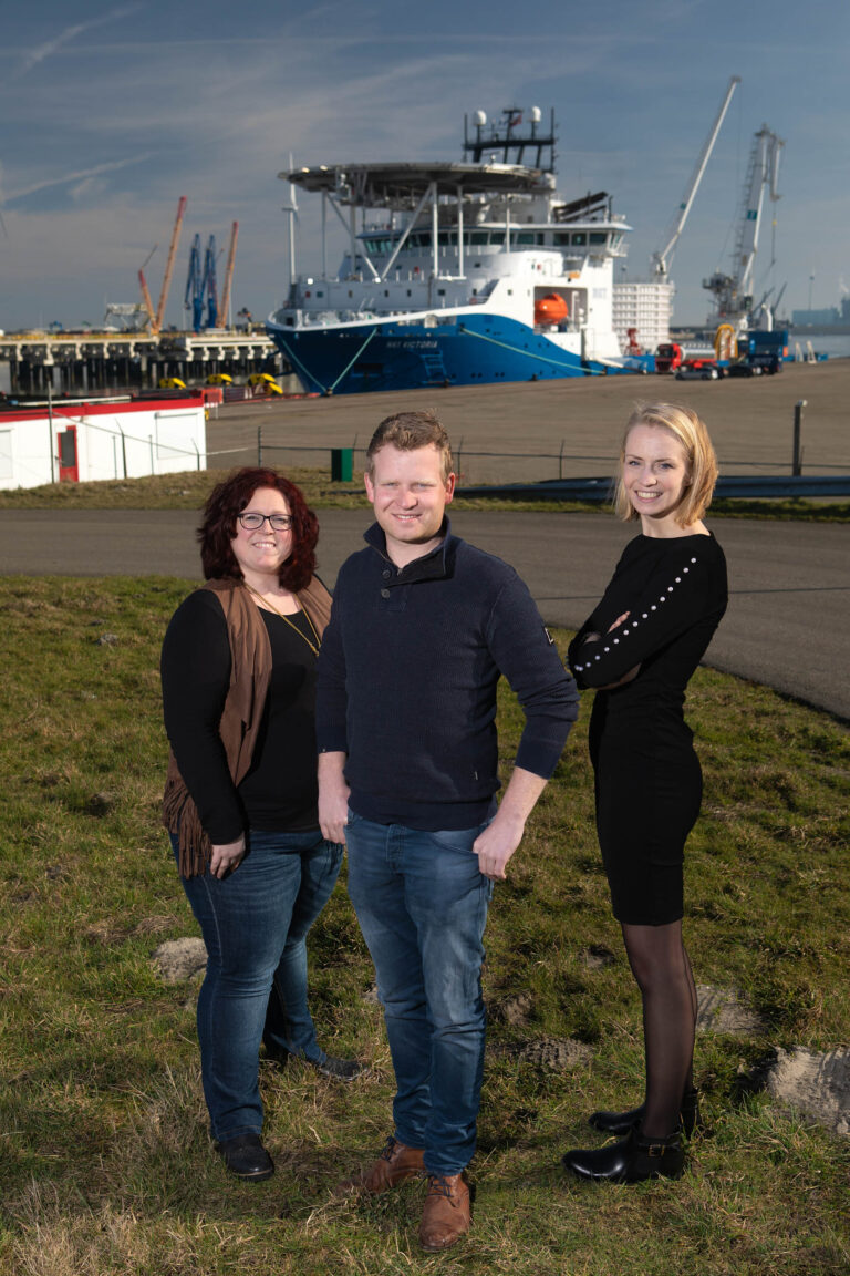 Deze foto, gemaakt door Jan Buwalda in Groningen, toont drie personen die buiten staan op een grasveld met een grote haven in de achtergrond. Op de achtergrond is een groot schip zichtbaar met kranen en verschillende schepen, wat duidt op een industriële havenomgeving. De lucht is helder, wat zorgt voor goede verlichting. De personen op de foto kijken naar de camera en zien er ontspannen en vriendelijk uit.