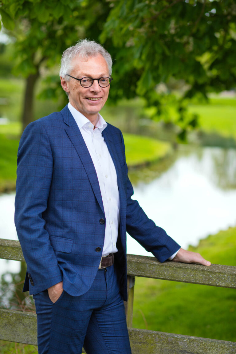 Dit is een portretfoto van een man in een formel blauwe ruitjesblazer en een witte overhemd. Hij draagt een bril en glimlacht ontspannen naar de camera, terwijl hij met één hand leunt op een houten hek. Op de achtergrond is een schilderachtig landschap te zien met groen gras en een rustige vijver, omringd door bomen. De foto straalt een professionele en vriendelijke sfeer uit. Fotograaf: Jan Buwalda, Groningen.