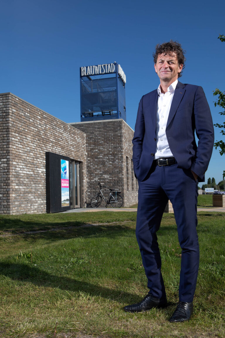 Deze foto, genomen door Jan Buwalda, toont een man in een donkerblauw pak die buiten staat, tegen de achtergrond van een modern gebouw met de tekst Blauwestad erop. De lucht is helderblauw en er staat een fiets tegen de muur. Het geheel straalt een professionele en zonnige sfeer uit.