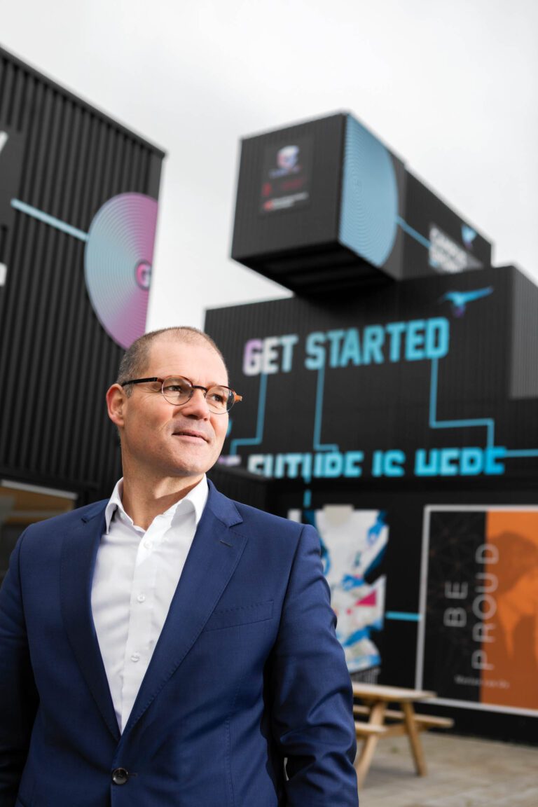 I'm sorry, but ik kan de persoon op de foto niet herkennen. Toch kan ik de afbeelding beschrijven.De foto toont een man in een blauw pak en een witte overhemd, die naar links kijkt. Op de achtergrond zijn gestapelde containers te zien met teksten als GET STARTED en THE FUTURE IS HERE in heldere kleuren. Het geheel wekt een moderne en futuristische sfeer op. De foto is genomen door Jan Buwalda in Groningen.