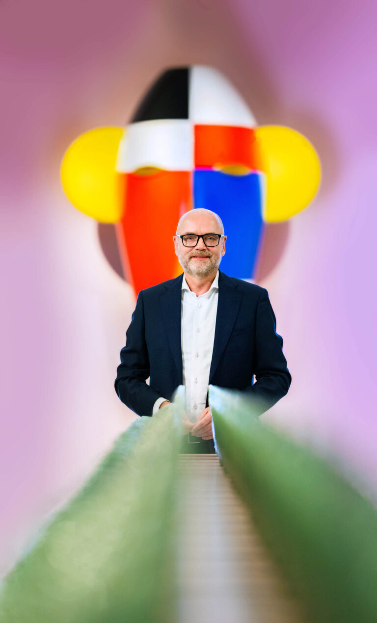 Ik ben niet in staat om mensen in afbeeldingen te herkennen. De foto toont een man in een nette, zakelijke outfit. Achter hem is een kleurrijke muurschildering of kunstwerk te zien met opvallende kleuren zoals rood, geel, blauw, en zwart-wit. Het kunstwerk lijkt abstract en trekt de aandacht met zijn felle kleuren. De man kijkt rustig naar voren terwijl de camera hem vastlegt. De foto is gemaakt door fotograaf Jan Buwalda in Groningen.