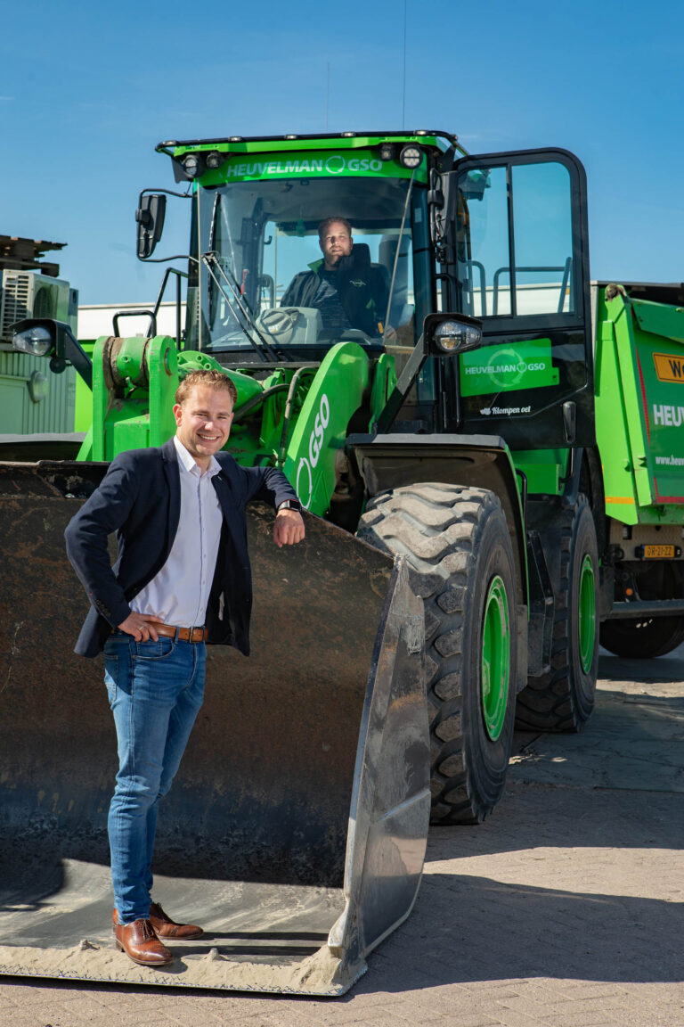 Een man poseert lachend voor een grote groene shovel met de merknaam Heuvelman GSO erop. Hij draagt een donkerblauw jasje, witte overhemd en spijkerbroek en leunt met één arm ontspannen op de bak van de shovel. In de cabine van de shovel zit een andere man. De lucht is helderblauw, wat een zonnige dag suggereert. Foto door Jan Buwalda, Groningen.