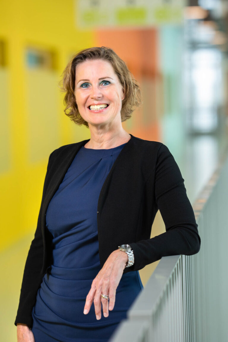 Het beeld toont een vrouw die glimlacht in een moderne, kleurrijke gang. Ze draagt een donkerblauwe jurk met een zwarte blazer en een zilveren horloge. Achter haar is een zachte, kleurrijke achtergrond zichtbaar, waarbij de kleuren geel en oranje opvallen. De foto is professioneel genomen en straalt een gevoel van vertrouwen en vriendelijkheid uit.Fotograaf: Jan Buwalda, Groningen.