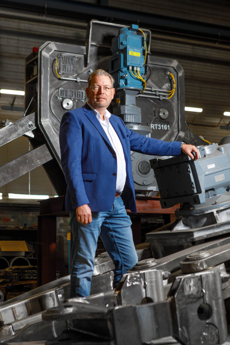 Dit is een foto van een man in een blauwe jas en jeans, staand in een industriële omgeving. Hij leunt tegen grote machineriedelen in een werkplaats of fabriekshal. De omgeving is goed verlicht en de focus ligt op de persoon en het metalen apparaat naast hem. Fotograaf: Jan Buwalda, Groningen.