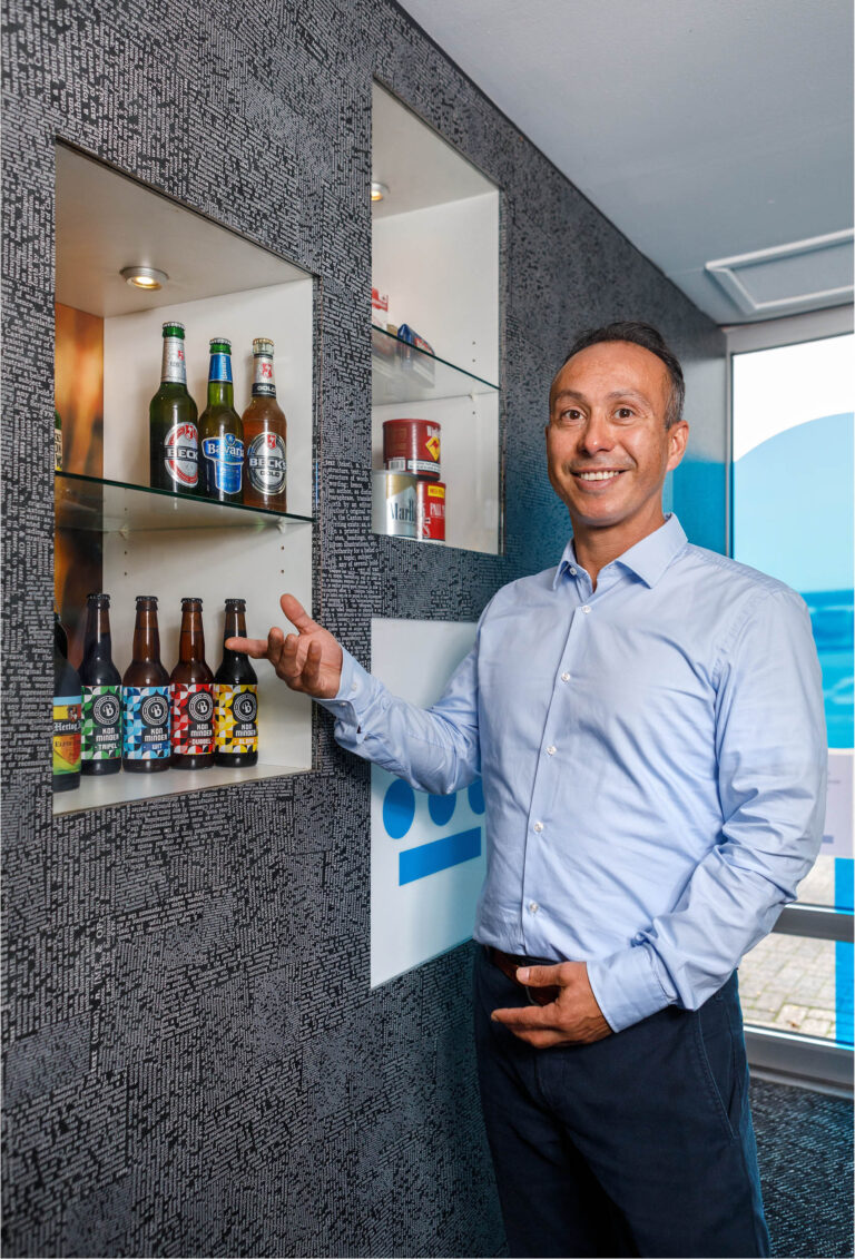 In deze foto, genomen door Jan Buwalda in Groningen, staat een man in een helder blauwe overhemd voor een wand met ingebouwde vitrines. De vitrines bevatten diverse bierflessen en blikjes, zorgvuldig uitgestald. De achtergrond heeft een modern patroon dat de aandacht vestigt op de drankverzameling. De man glimlacht naar de camera, wat een gastvrije en professionele sfeer creëert.