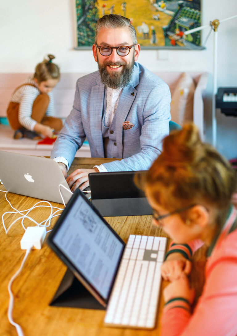 Dit beeld toont een man in een lichtblauw pak die glimlachend aan een tafel met verschillende elektronische apparaten werkt. Hij lijkt thuis te zijn, met kinderen op de achtergrond bezig met hun eigen activiteiten. De warme en huiselijke sfeer wordt benadrukt door de heldere kleuren en de kunst aan de muur. Fotografie door Jan Buwalda, Groningen.