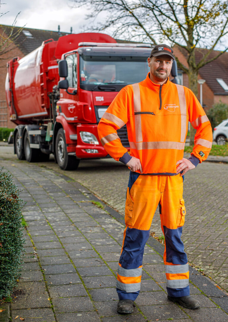 Een man gekleed in een feloranje en blauwe veiligheidsoutfit staat trots voor een grote rode vuilniswagen. Hij draagt een oranje trui met reflecterende strepen en een pet. De achtergrond toont een rustige straat met enkele huizen en bomen. De foto straalt een gevoel van dagelijks werkende mensen uit die een belangrijke rol spelen in de gemeenschap. Deze foto is gemaakt door Jan Buwalda in Groningen.