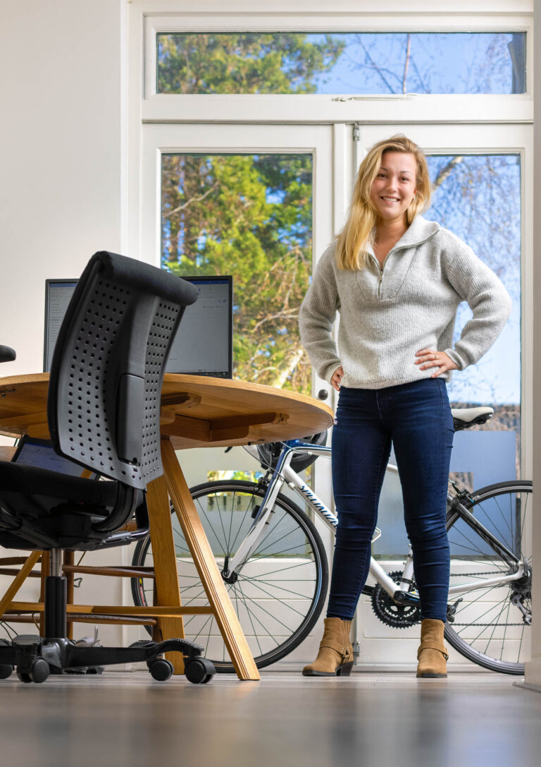 Ik kan je zeggen dat de foto een persoon toont die binnen in een ruimte staat bij een houten tafel met computers, met een fiets op de achtergrond. Het lijkt een zonnige dag te zijn, gezien het licht dat door het raam valt. Helaas ken ik de details van de persoon niet. Deze foto is genomen door fotograaf Jan Buwalda in Groningen.