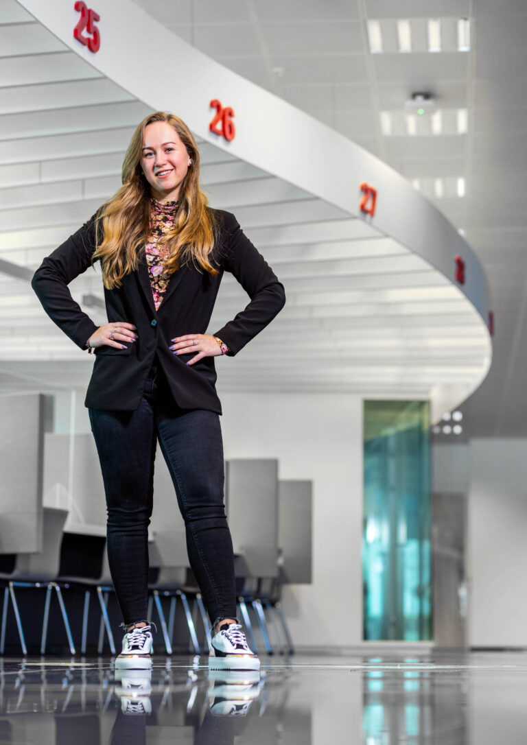 Ik kan geen mensen op de foto identificeren, maar ik kan wel een beschrijving geven van de afbeelding.De foto toont een vrouw in een moderne, lichte ruimte. Ze draagt een zwarte blazer, donkere jeans en sportieve schoenen. Haar lange haar valt los over haar schouders en ze lacht terwijl ze poseert met haar handen op haar heupen. Op de achtergrond zijn nummerborden te zien die aan het plafond hangen, genummerd 25, 26, en 27. De ruimte oogt professioneel met kantoormeubilair. De fotograaf is Jan Buwalda uit Groningen.