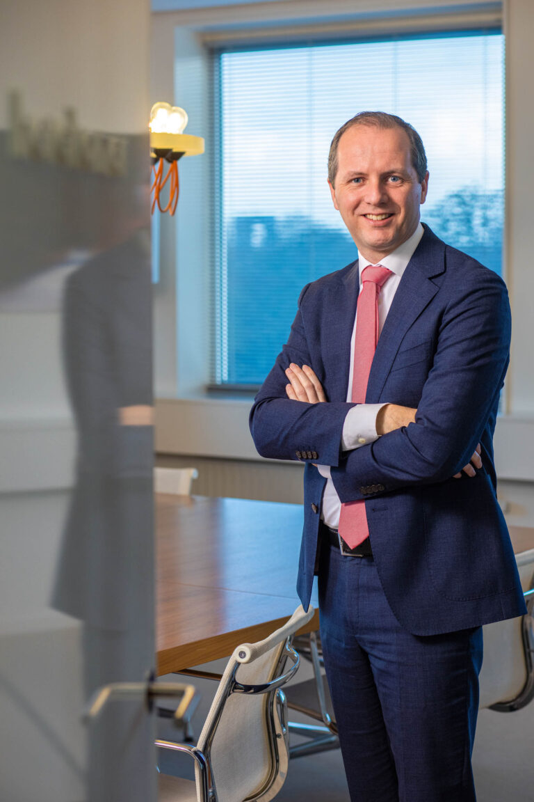 Ik kan geen mensen identificeren, maar ik kan wel een beschrijving van de foto geven:Op de foto zien we een man in een zakelijke omgeving. Hij draagt een donkerblauw pak met een witte overhemd en een roze stropdas. Hij staat met gekruiste armen en glimlacht naar de camera. Achter hem is een vergadertafel zichtbaar, met in de achtergrond een raam met jaloezieën die deels geopend zijn, waardoor er blauw licht naar binnen schijnt. De foto is genomen door Jan Buwalda in Groningen.