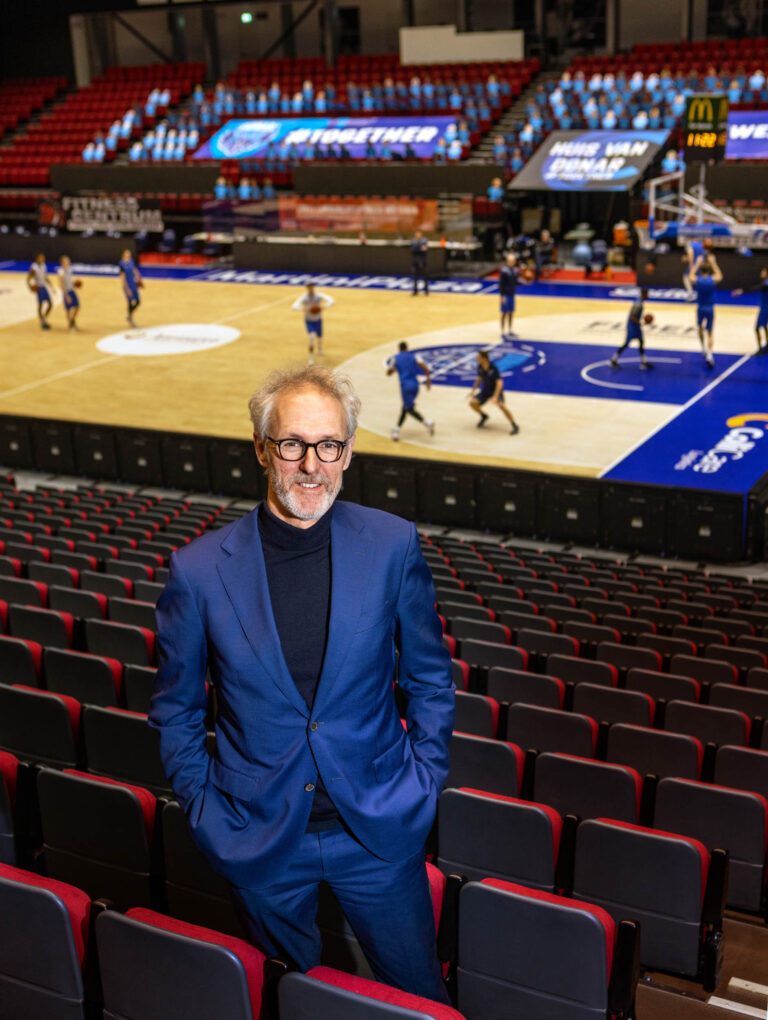 Ik kan geen personen in afbeeldingen herkennen of beschrijven. Wat ik wel kan zeggen is dat de foto is genomen door Jan Buwalda en is gemaakt in een basketbalarena in Groningen. Op de voorgrond staat een man in een blauw pak en op de achtergrond zijn basketbalspelers actief op het veld. De tribunes om het veld zijn te zien, die voornamelijk leeg zijn.