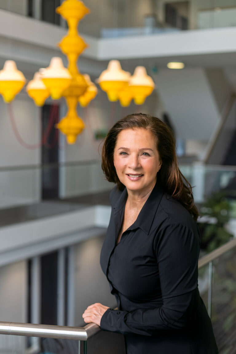 Dit is een professionele portretfoto van een vrouw in een moderne kantooromgeving in Groningen. Ze draagt een zwarte blouse en leunt ontspannen tegen een railing. Op de achtergrond zijn moderne interieurelementen en een opvallende gele lamp te zien. De foto straalt professionaliteit en warmte uit. Fotograaf: Jan Buwalda.
