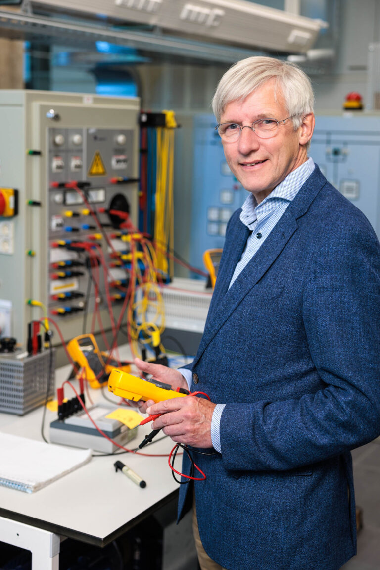 Het beeld toont een man in een laboratoriumomgeving met diverse technische apparatuur. Hij draagt een donkerblauw colbert en een lichtblauw overhemd. De man houdt meetapparatuur in zijn handen en kijkt vriendelijk lachend in de camera. Op de achtergrond zijn schakelkasten en kabels zichtbaar, wat de technologische setting benadrukt. Foto door Jan Buwalda, Groningen.
