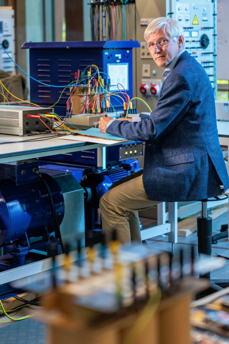 Ik kan de persoon op de foto niet identificeren, maar hier is een beschrijving:In een moderne technische omgeving zit een wat oudere man aan een werkbank. Hij werkt geconcentreerd met verschillende elektrische apparatuur, waarbij kleurrijke kabels verbonden zijn met een meetinstrument. De ruimte om hem heen is gevuld met geavanceerde machines en technische instrumenten, wat de indruk geeft van een laboratorium of werkplaats voor elektronica. De sfeer is professioneel en innovatief.Fotograaf: Jan Buwalda, Groningen.