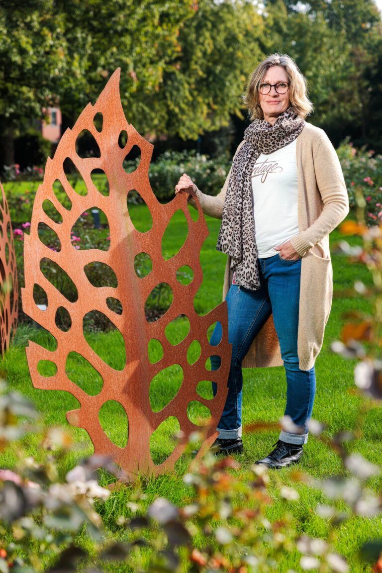 De foto toont een vrouw staande in een groene tuin, leunend tegen een decoratief metalen kunstwerk met een uitgesneden organisch bladpatroon. Ze draagt een casual outfit met een beige vest, jeans, en een sjaal met luipaardprint. Het kunstwerk en de omgeving stralen een gevoel van herfst uit, met bomen en bloemen op de achtergrond. Foto door Jan Buwalda in Groningen.