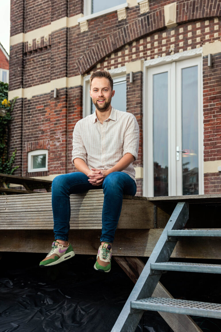 Dit is een portretfoto gemaakt door Jan Buwalda. Een man zit ontspannen op een houten platform buiten voor een bakstenen gebouw met een grote glazen deur. Hij draagt een lichtgekleurde overhemd en een spijkerbroek, en zijn groene sneakers voegen een vleugje kleur toe aan de uitstraling. Op de achtergrond is de karakteristieke architectuur van Groningen te zien.