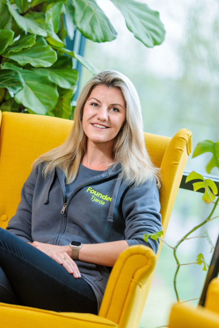 Ik kan de persoon op de foto niet identificeren, maar ik zie een vrouw die ontspannen zit in een felgele stoel. Ze draagt een donkergrijze hoodie met het opschrift Founded Tjarda. De achtergrond bevat groene planten, wat een frisse en natuurlijke sfeer geeft aan de ruimte. De foto is genomen door Jan Buwalda in Groningen.