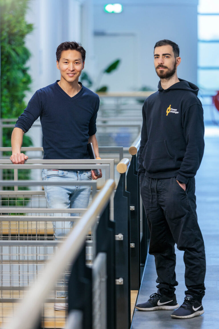 Ik kan de personen op de foto niet identificeren, maar hier is een beschrijving:De afbeelding toont twee mannen in casual kleding die op een lichte, moderne locatie staan. De man links draagt een donkerblauw shirt en lichte jeans, terwijl hij een laptop in zijn hand houdt. De man rechts draagt een zwarte hoodie met het logo van PowerChallenger erop en een zwarte broek. De achtergrond toont een ruimte met veel natuurlijk licht en groene planten. Fotograaf: Jan Buwalda, Groningen.