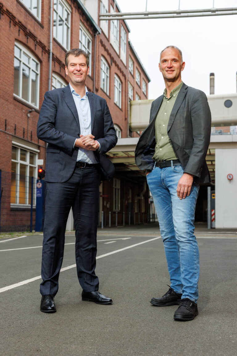 Twee mannen poseren buiten voor een gebouw met rode bakstenen en grote ramen. Ze dragen nette kleding; de ene man heeft een donker pak aan en de ander een colbert met spijkerbroek. De omgeving lijkt stedelijk en professioneel. Foto gemaakt door Jan Buwalda in Groningen.