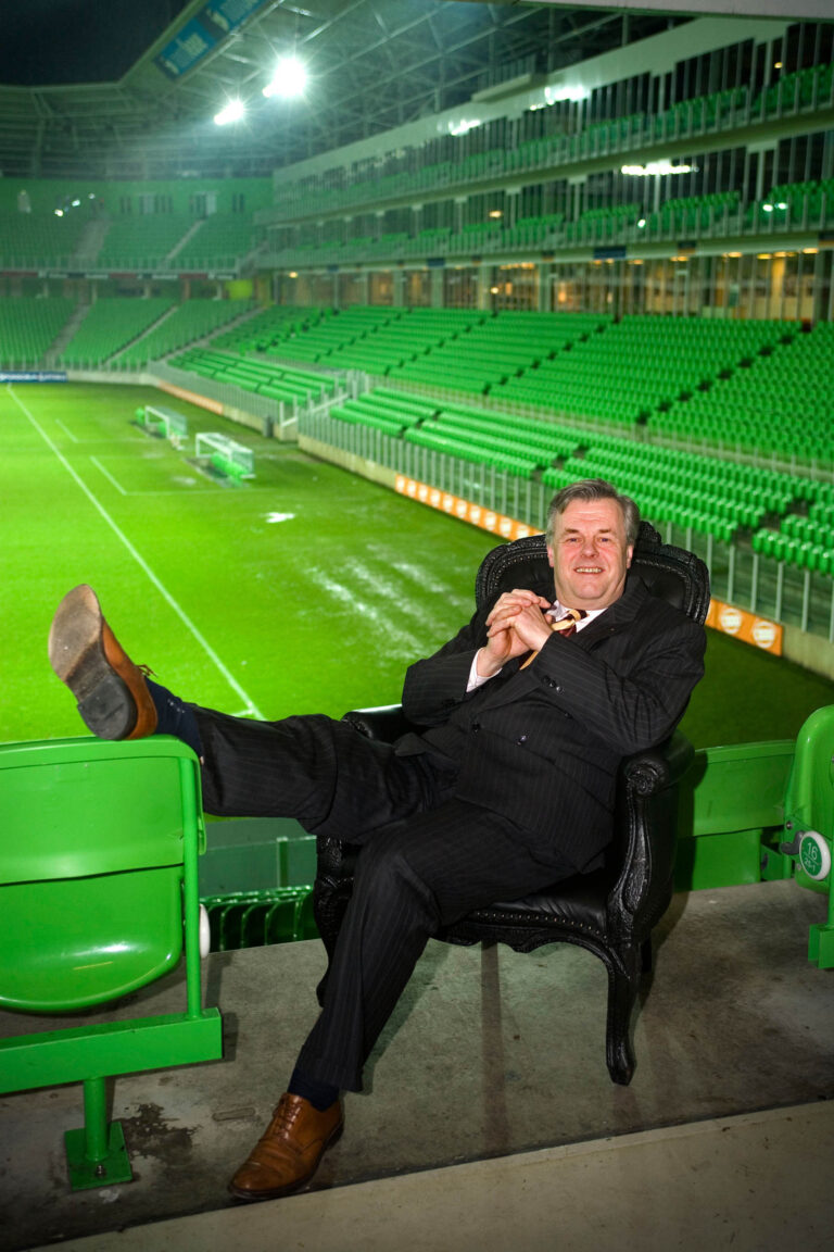 Ik ben niet in staat om personen te herkennen in foto's, maar ik kan wel een beschrijving maken van de foto.In het beeld zien we een man in een donker pak, ontspannen zittend op een sierlijke zwarte stoel, met een been over de rand van een tribune in een voetbalstadion. Het stadion is leeg en de tribunes zijn felgroen, wat een sterke visuele indruk geeft. De verlichting van het stadion geeft een heldere gloed aan de scene. De foto straalt een gevoel van rust en comfort uit in wat normaal een drukke en levendige omgeving zou zijn. Foto door Jan Buwalda in Groningen.