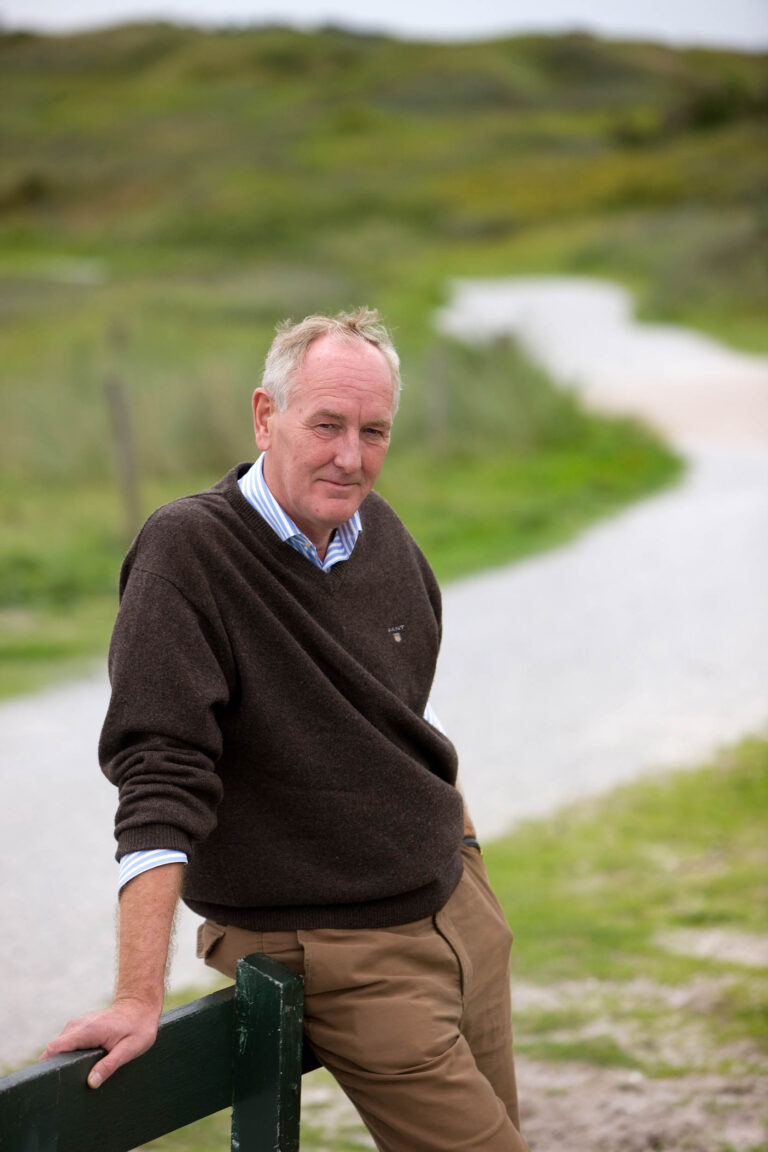 Een man leunt ontspannen tegen een hek, gekleed in een bruine trui en beige broek, terwijl op de achtergrond een kronkelend pad en groene velden in beeld zijn. De omgeving straalt een rustige en landelijke sfeer uit. Foto door Jan Buwalda, Groningen.