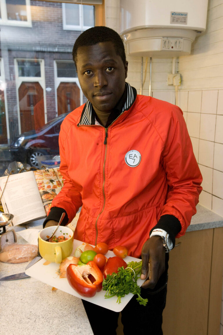 Deze foto toont een persoon in een keuken in Groningen, die trots een snijplank met verse ingrediënten vasthoudt. Hij draagt een opvallend rode jas en naast hem ligt een opengeslagen kookboek. De keuken heeft een raam dat uitkijkt op een rij huizen buiten. Fotograaf: Jan Buwalda.