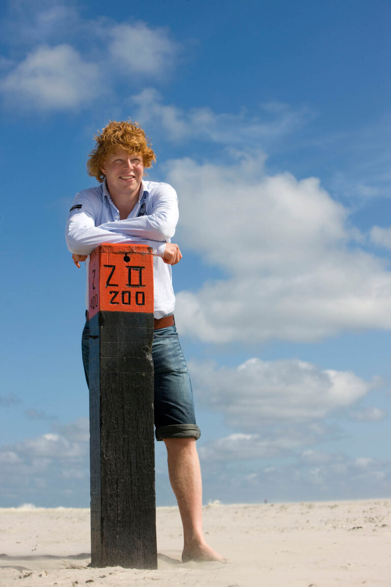 Ik kan geen mensen op de foto herkennen, maar ik kan wel een beschrijving geven van de scène.Op de foto staat een persoon die leunt tegen een zwart-rood paaltje in een zandlandschap. Het is een zonnige dag met een heldere blauwe lucht en enkele wolken. De persoon draagt een licht overhemd met opgerolde mouwen en een spijkerbroek, die ook is opgerold tot halverwege de kuiten. De foto straalt een ontspannen en zomers gevoel uit.Fotograaf: Jan Buwalda uit Groningen.