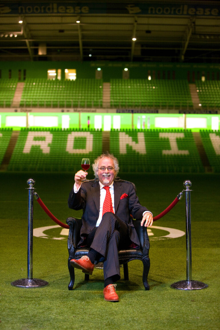 Een man in een gestreept pak en rode schoenen zit ontspannen op een elegante stoel geplaatst op het voetbalveld in het stadion van FC Groningen. Hij heft een glas wijn, glimlachend naar de camera. Het stadion op de achtergrond is herkenbaar aan de groene stoelen en de grote letters die GRONINGEN spellen. De setting geeft een gevoel van luxe en exclusiviteit. Fotografie door Jan Buwalda.