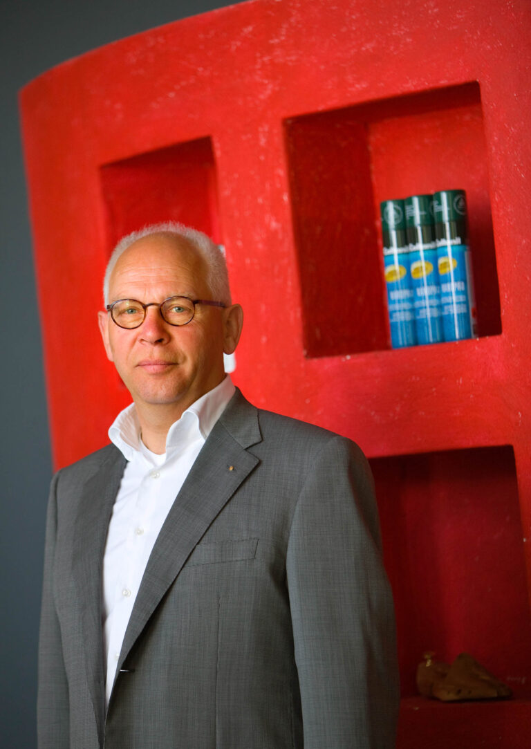 Het beeld toont een man in een grijs pak met een witte overhemd. Hij draagt een bril en kijkt zelfverzekerd naar de camera. Op de achtergrond is een rode muur met enkele producten zichtbaar. Deze foto is genomen door Jan Buwalda in Groningen.