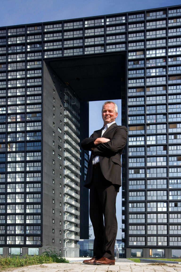 Ik kan deze persoon niet identificeren, maar hier is een beschrijving van de foto:Een man in een donker pak met een witte blouse staat met zijn armen over elkaar voor een modern kantoorgebouw in Groningen. Het gebouw heeft een opvallende architectuur met een centrale open ruimte in de vorm van een rechthoek. De lucht is helder en blauw, wat de indrukwekkende hoogte van de structuur accentueert. Deze foto is gemaakt door Jan Buwalda.