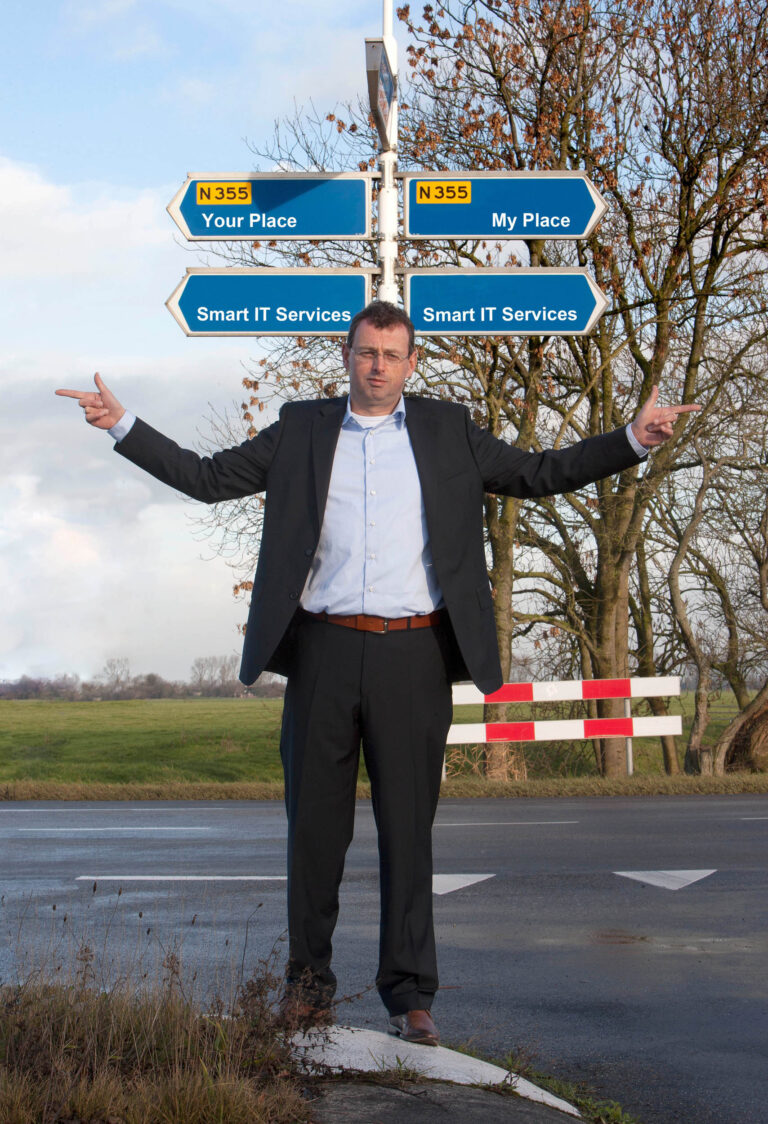 Een man in een net pak staat op een kruispunt voor een wegwijzer met daarop teksten als Your Place en My Place onder een blauwe lucht. Hij wijst met beide handen naar tegengestelde richtingen. De omgeving is landelijk, met grasvelden en bomen zonder bladeren. De foto is gemaakt door Jan Buwalda in Groningen.