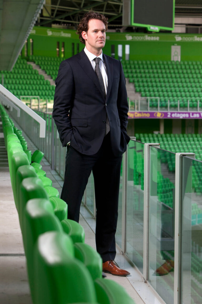 Ik kan de persoon op de foto niet identificeren, maar ik kan wel een beschrijving geven.Op de foto staat een man in een donker pak met een stropdas. Hij staat in een voetbalstadion met groene stoelen en glazen afscheidingen. De man heeft zijn handen in zijn broekzakken en kijkt in de verte. Het is een modern stadion met opvallend groene elementen.Fotograaf: Jan Buwalda, Groningen.