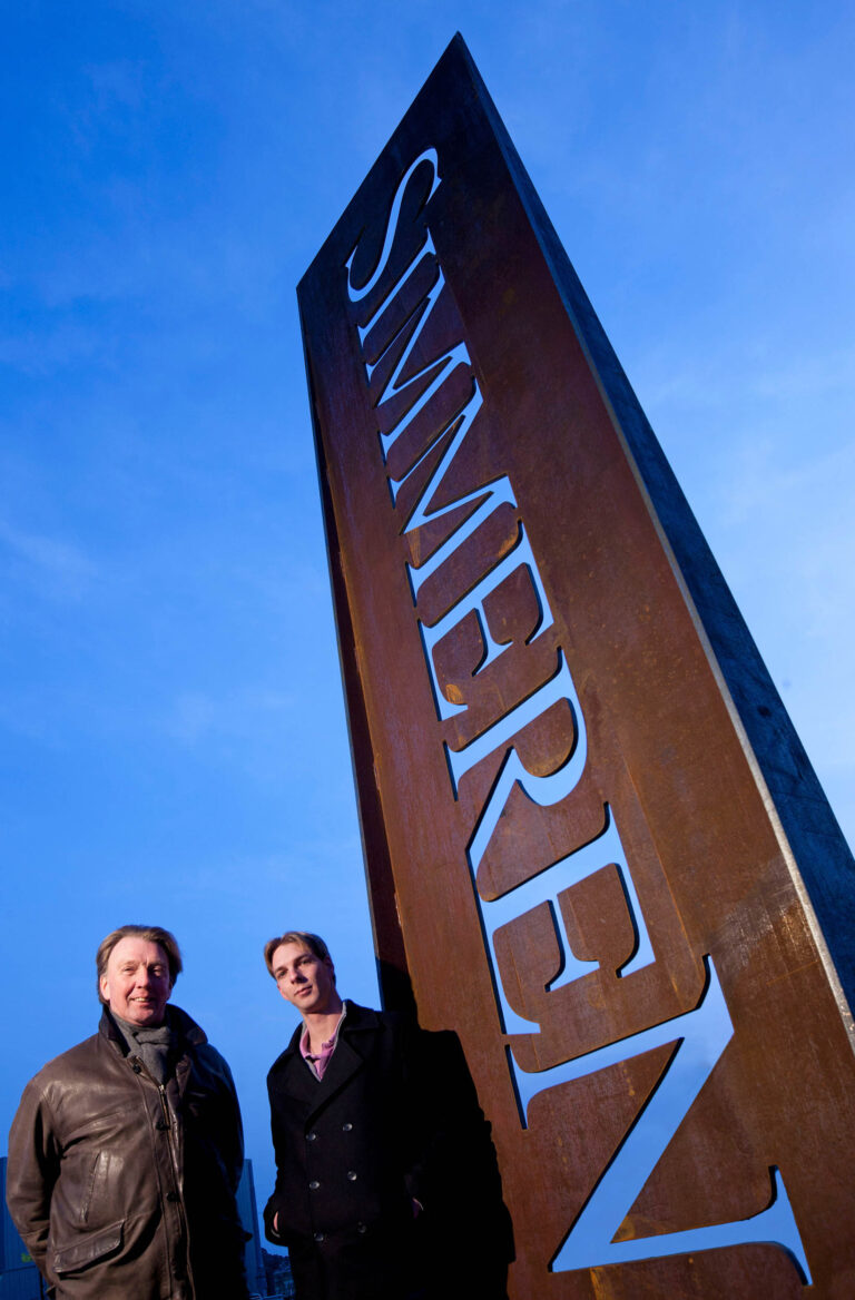 Dit is een foto van twee mannen die voor een groot metalen kunstwerk staan met de tekst Simmeren erop gegraveerd. De lucht is helder blauw en de belichting van de foto zorgt voor een opvallend contrast tussen de achtergrond en het kunstwerk. De mannen zijn gekleed in donkere jassen. Deze foto is genomen door Jan Buwalda in Groningen.