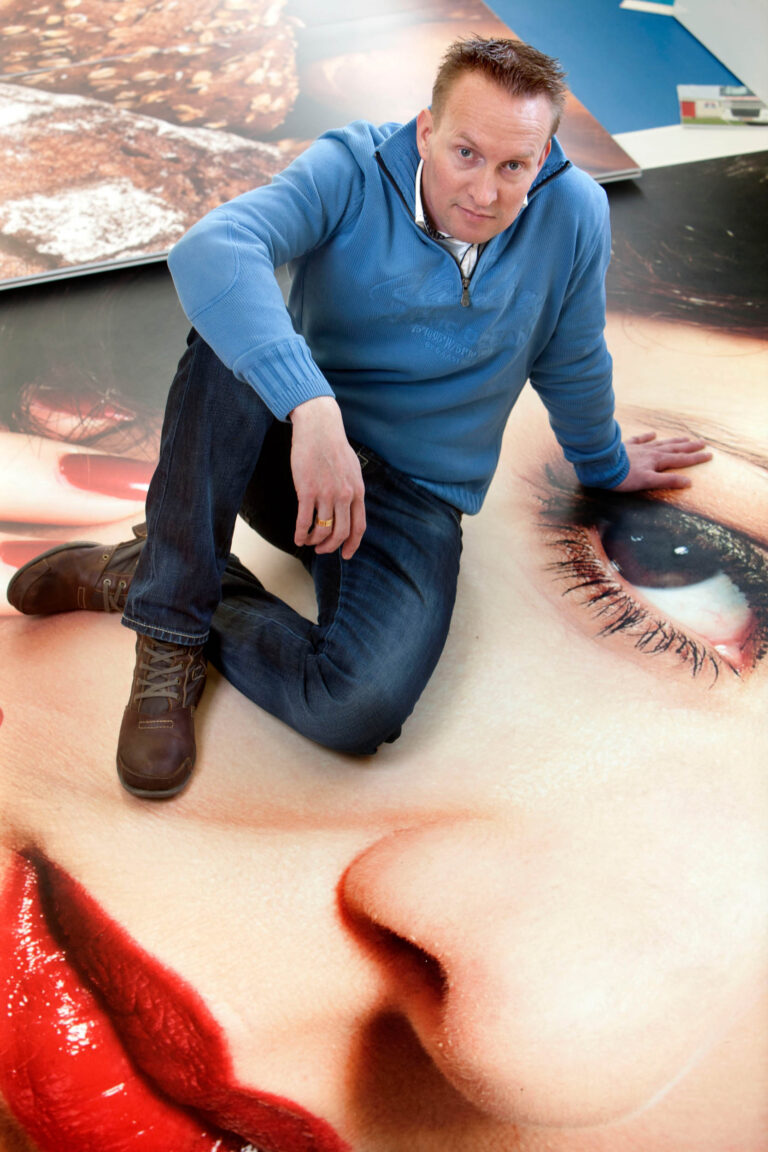 Het beeld toont een man in een blauwe trui en jeans, gehurkt op een grote afbeelding van een gezicht met rode lippen. Hij kijkt direct in de camera. Er zijn andere grote afdrukken zichtbaar op de achtergrond. Foto door Jan Buwalda, Groningen.