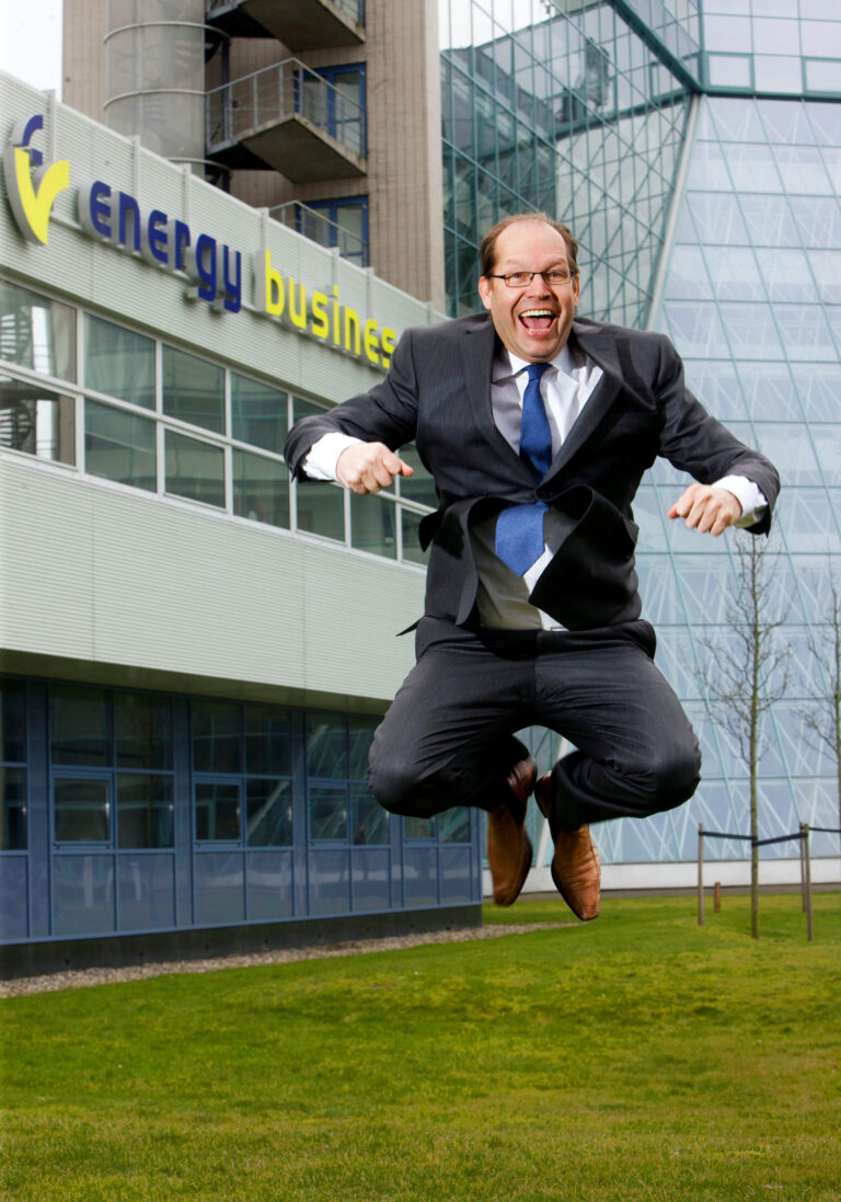 Ik kan geen mensen identificeren, maar ik kan wel een beschrijving van de foto geven.Op de foto is een man te zien die in een spontane sprong is vastgelegd. Hij draagt een donker pak met een witte overhemd en een blauwe stropdas. De man toont een energieke en blije uitdrukking. Op de achtergrond is een modern kantoorgebouw zichtbaar met daarop de tekst energy business. De foto is genomen op een groene grasvlakte voor het gebouw.Fotograaf: Jan Buwalda, Groningen.