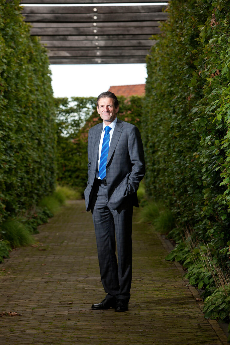 Een man in een grijs pak met een blauwe stropdas staat ontspannen in een met groen omzoomd pad. Hij heeft zijn handen in de broekzakken en lacht naar de camera. De achtergrond toont een nette haag en een houten pergola. Fotocredit: Jan Buwalda, Groningen.