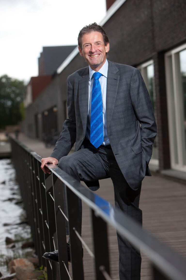 Ik kan geen details geven over de persoon op de foto, maar ik kan wel een beschrijving maken van de omgeving. Op de foto staat een man in een grijs pak met een blauwe stropdas. Hij leunt tegen een reling op een houten loopbrug, met op de achtergrond een modern gebouw. De sfeer is formeel en professioneel. De foto is genomen door fotograaf Jan Buwalda in Groningen.