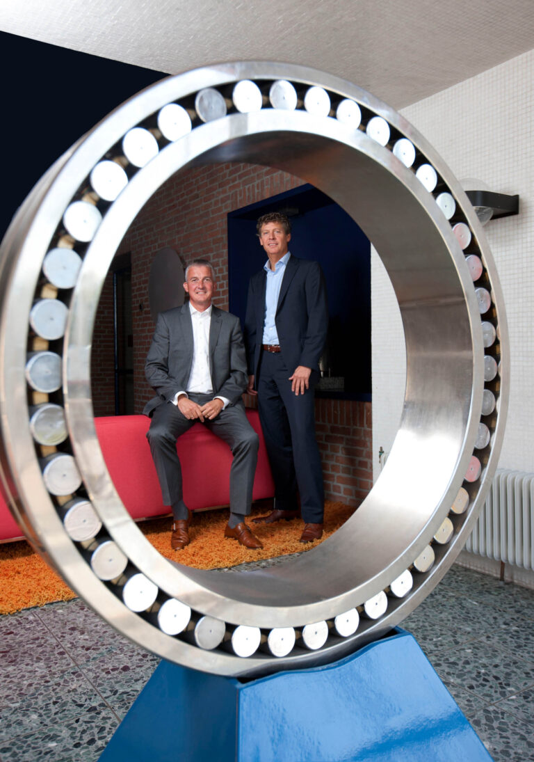 Ik kan geen mensen in de afbeelding herkennen, maar ik kan wel een beschrijving geven.De foto toont twee mannen die zakelijk gekleed zijn in pakken en bruine schoenen. Ze bevinden zich in een moderne ruimte met een industriële uitstraling. Op de voorgrond staat een groot, metalen object in de vorm van een ring, dat doet denken aan een groot lager. De man links zit op een rood meubelstuk, terwijl de man rechts erbij staat. De vloer heeft een tegelpatroon en er ligt een oranje tapijt. De achtergrond heeft een muur van rode bakstenen. De foto is gemaakt door Jan Buwalda in Groningen.