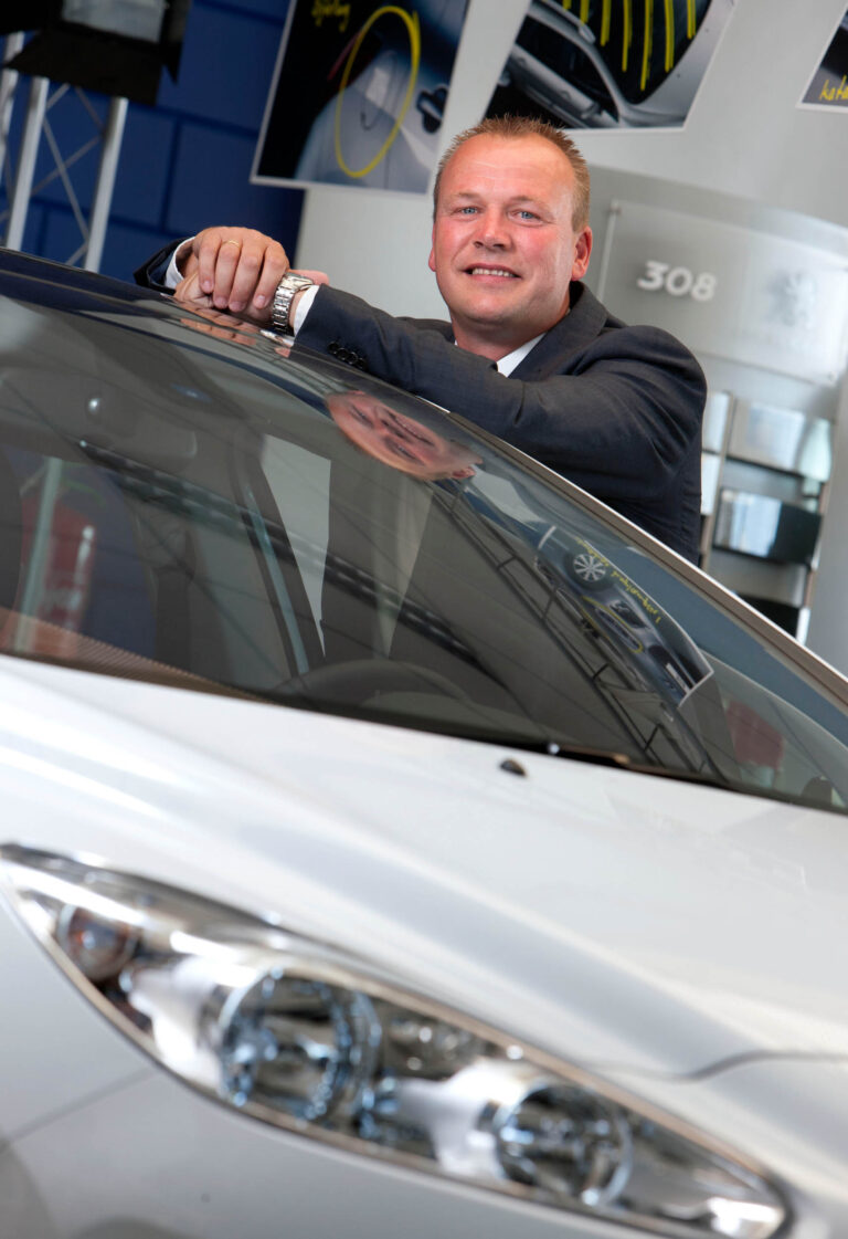 Dit is een foto van een man die ontspannen tegen een auto leunt en glimlacht naar de camera. De achtergrond toont een moderne autodealeromgeving met afbeeldingen van auto's aan de muur. De auto op de voorgrond heeft een witte carrosserie. Fotograaf: Jan Buwalda, Groningen.