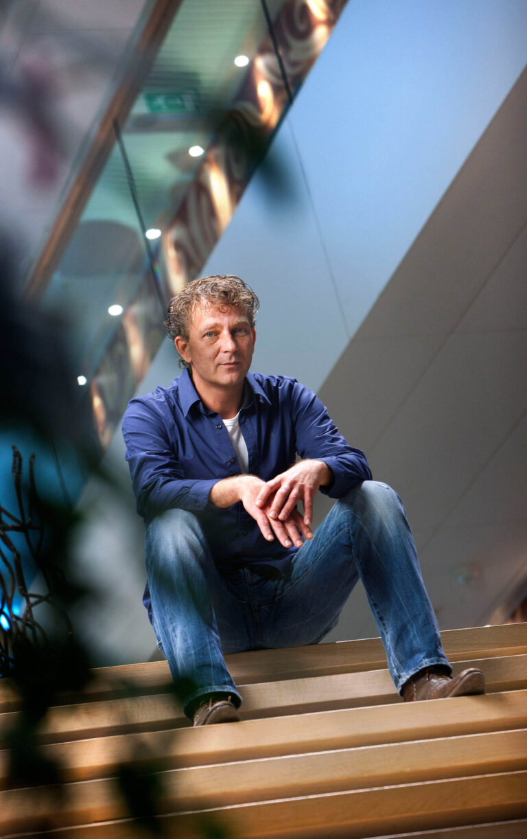 Deze foto toont een persoon die ontspannen zit op een trap in een moderne, lichte ruimte. De persoon draagt een donkerblauw overhemd en jeans, en kijkt recht in de camera. Het interieur heeft een strakke architectuur met glazen en metalen elementen. De foto is gemaakt door Jan Buwalda in Groningen.
