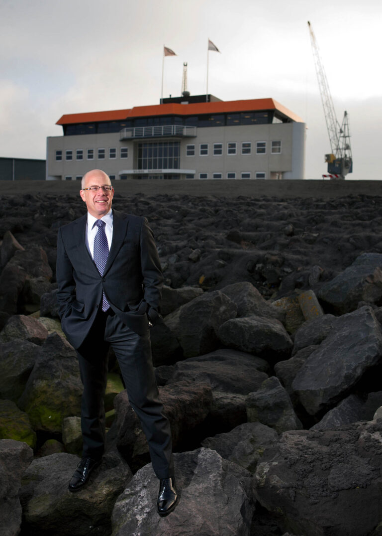 Ik kan de identiteit van mensen op foto's niet herkennen, maar ik kan wel de foto beschrijven. Op de foto staat een persoon in een net pak en das, poserend op een rotsachtig terrein. Op de achtergrond staat een modern gebouw met grote ramen en een oranje dak. Er is ook een kraan zichtbaar aan de rechterkant. De lucht is bewolkt. De foto lijkt professioneel te zijn genomen vanuit een laag camerastandpunt, wat de persoon prominent in beeld brengt ten opzichte van de achtergrond. Fotograaf: Jan Buwalda, Groningen.