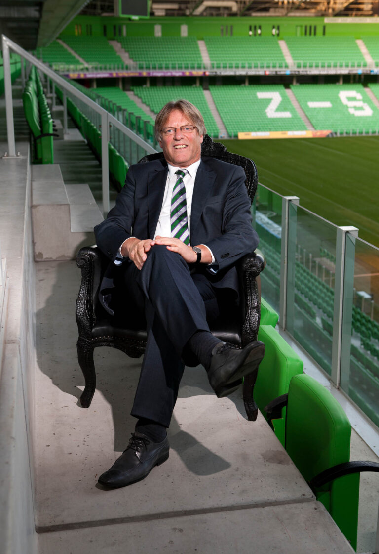 Het beeld toont een man in een pak met een stropdas, zittend op een sierlijke zwarte stoel in een bijna leeg voetbalstadion. De stoelen in het stadion zijn groen, met de letters Z-S zichtbaar op de achtergrond. De man kijkt ontspannen en glimlachend naar de camera. De foto is genomen door Jan Buwalda in Groningen.
