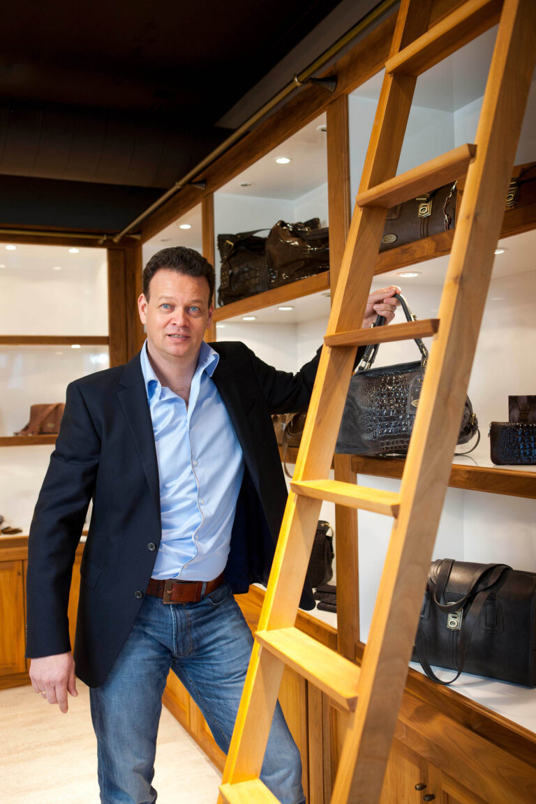 Dit is een foto van een man die in een winkel staat naast een houten ladder. Hij draagt een blauw jasje, een lichtblauw overhemd en een spijkerbroek. Op de achtergrond zijn leren tassen en accessoires te zien in een houten vitrinekast. De sfeer is professioneel en de winkel lijkt gespecialiseerd te zijn in leren goederen. Fotografie door Jan Buwalda, Groningen.