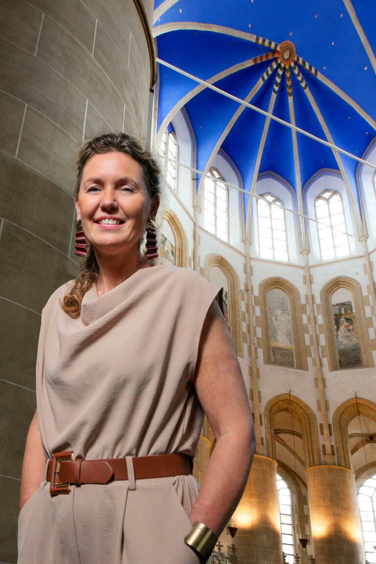 Ik kan de persoon op de foto niet identificeren, maar ik kan wel een beschrijving geven van de afbeelding.Op de foto staat een lachende vrouw in een beige outfit met een bruine riem en opvallende sieraden. Ze poseert in een imposant gebouw met hoge, versierde plafonds en grote glas-in-loodramen. De blauwe bekleding van het plafond vormt een mooi contrast met de beige en bruine tinten van het interieur. De foto is gemaakt door Jan Buwalda in Groningen.