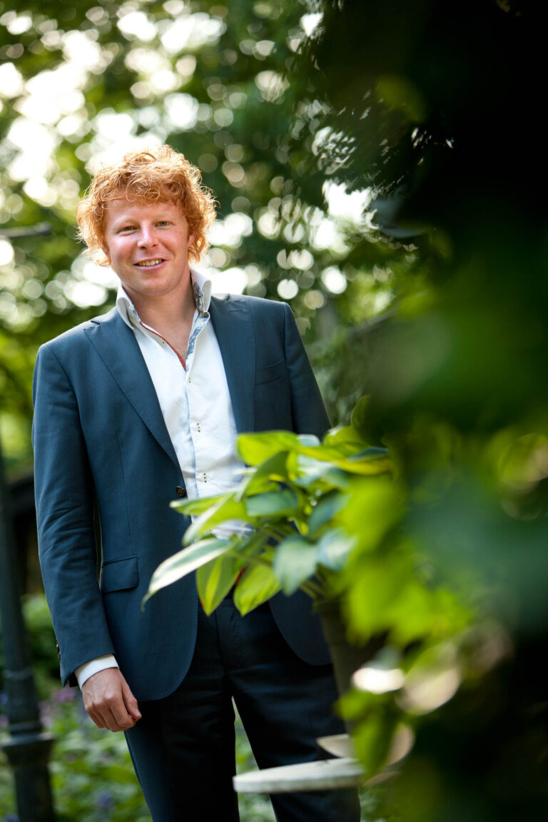 Het portret toont een man in een nette, donkerblauwe blazer en een wit overhemd. Hij staat buiten, omgeven door groene bladeren en de zon filtert zachtjes door de bomen, wat zorgt voor een bokeh-effect in de achtergrond. De sfeer is ontspannen en natuurlijk. Foto door Jan Buwalda in Groningen.