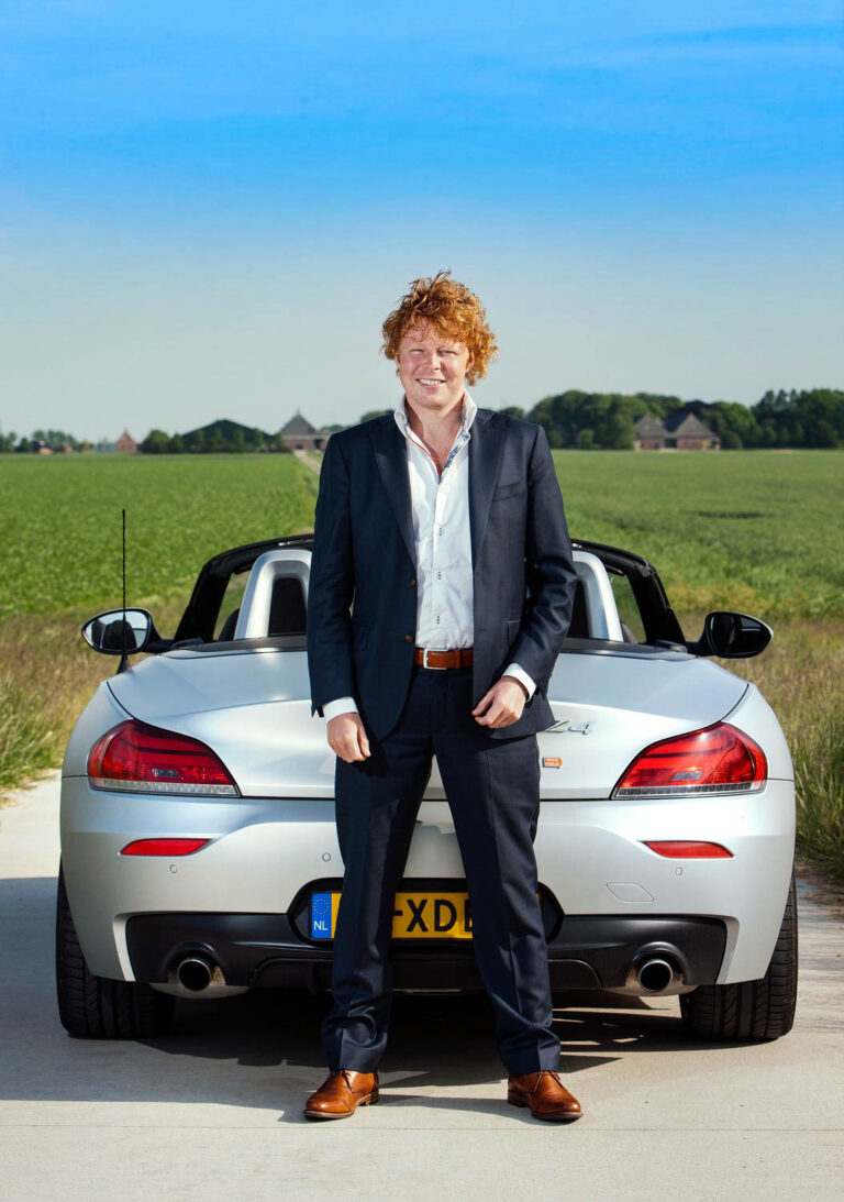 Een jonge man in een donkerblauw pak staat voor een zilverkleurige sportauto op een landelijke weg met groene velden en een heldere blauwe lucht in de achtergrond. De man heeft een open houding en draagt bruine schoenen, die elegant passen bij zijn stijl. Het is een zonnige dag en zowel de auto als het landschap stralen een gevoel van vrijheid en luxe uit. De foto is genomen door Jan Buwalda in Groningen.