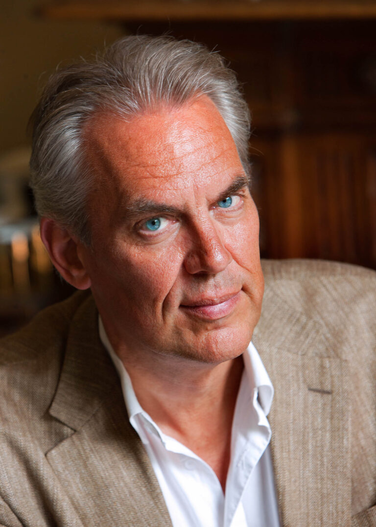 Het portret toont een man met grijs haar en blauwe ogen, gekleed in een lichtbruin colbert en een wit overhemd. Hij kijkt rechtstreeks in de camera met een serieuze maar warme blik. De achtergrond is onscherp, waardoor de focus volledig op het gezicht ligt. Deze foto is genomen door fotograaf Jan Buwalda uit Groningen.