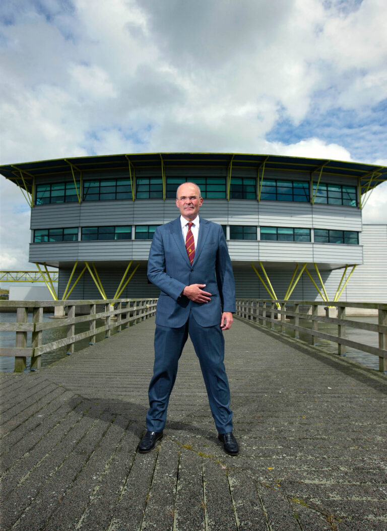 Een man in een blauw pak staat zelfverzekerd op een betonnen wandelpad met houten railing, dat naar een modern gebouw met een innovatieve architectuur leidt. Het gebouw heeft een metallic uitstraling met grote ramen en gele accenten bij de steunbalken. De lucht is grotendeels bewolkt, maar er zijn enkele stukken met blauwe lucht zichtbaar. De foto is genomen door Jan Buwalda in Groningen.