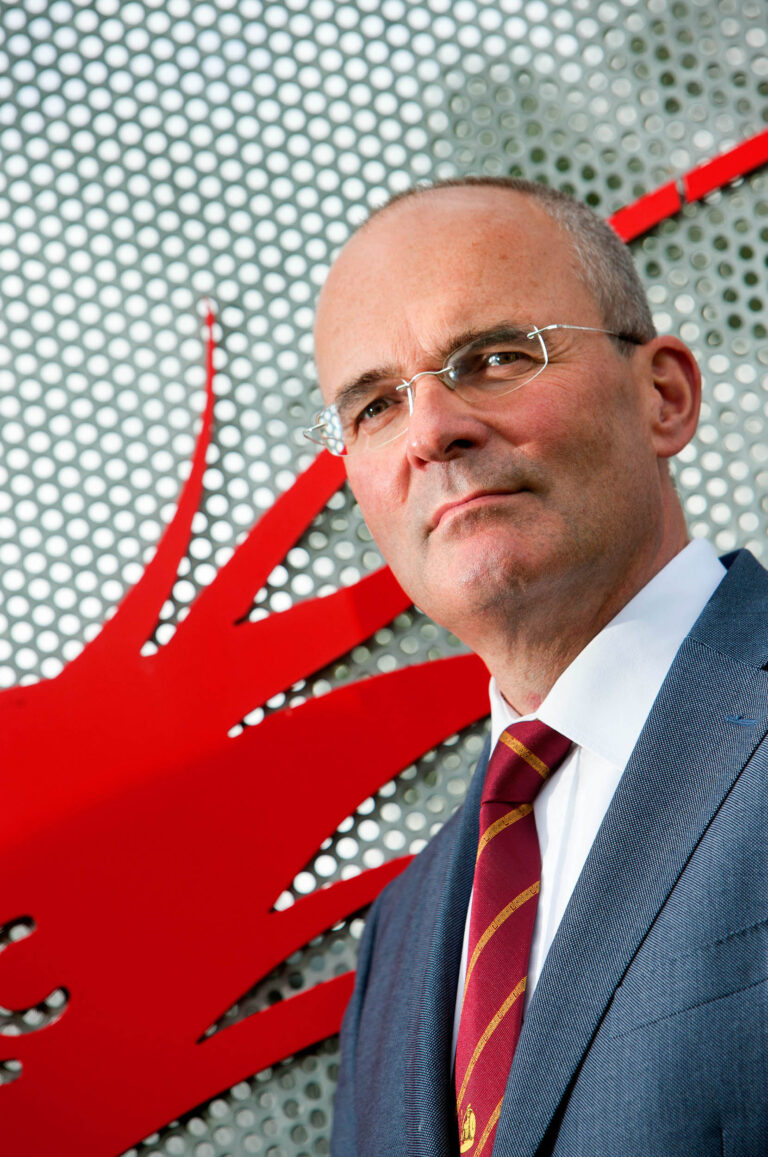 Dit is een portretfoto van een man in een net pak en stropdas. Hij draagt een bril en staat voor een achtergrond met een opvallend rood patroon en een grijze, geperforeerde structuur. De foto straalt een professionele en serieuze uitstraling uit. Fotograaf: Jan Buwalda uit Groningen.